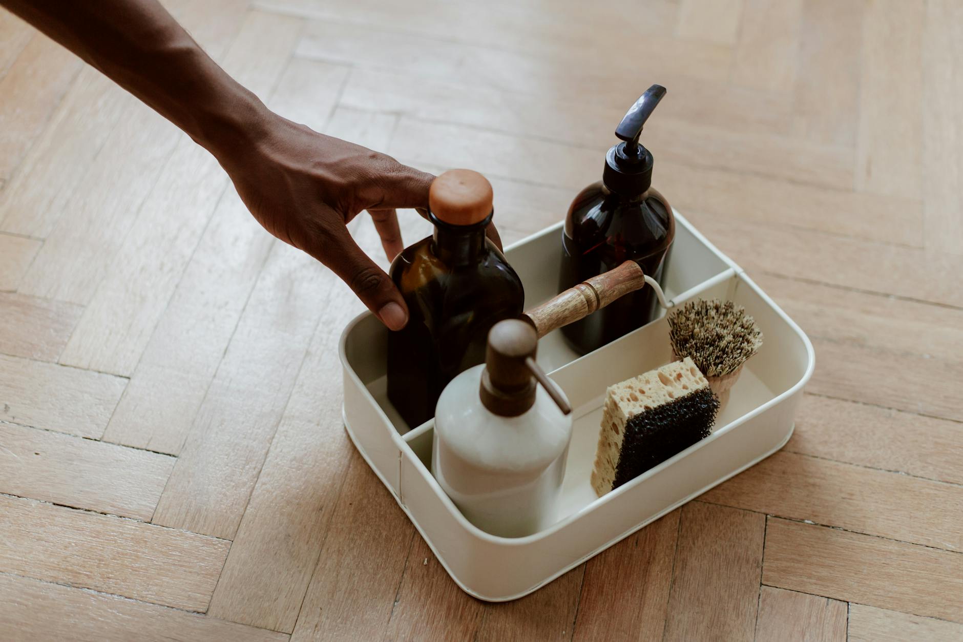 A hand reaching for a cleaning set with bottles and brushes on a wooden floor.